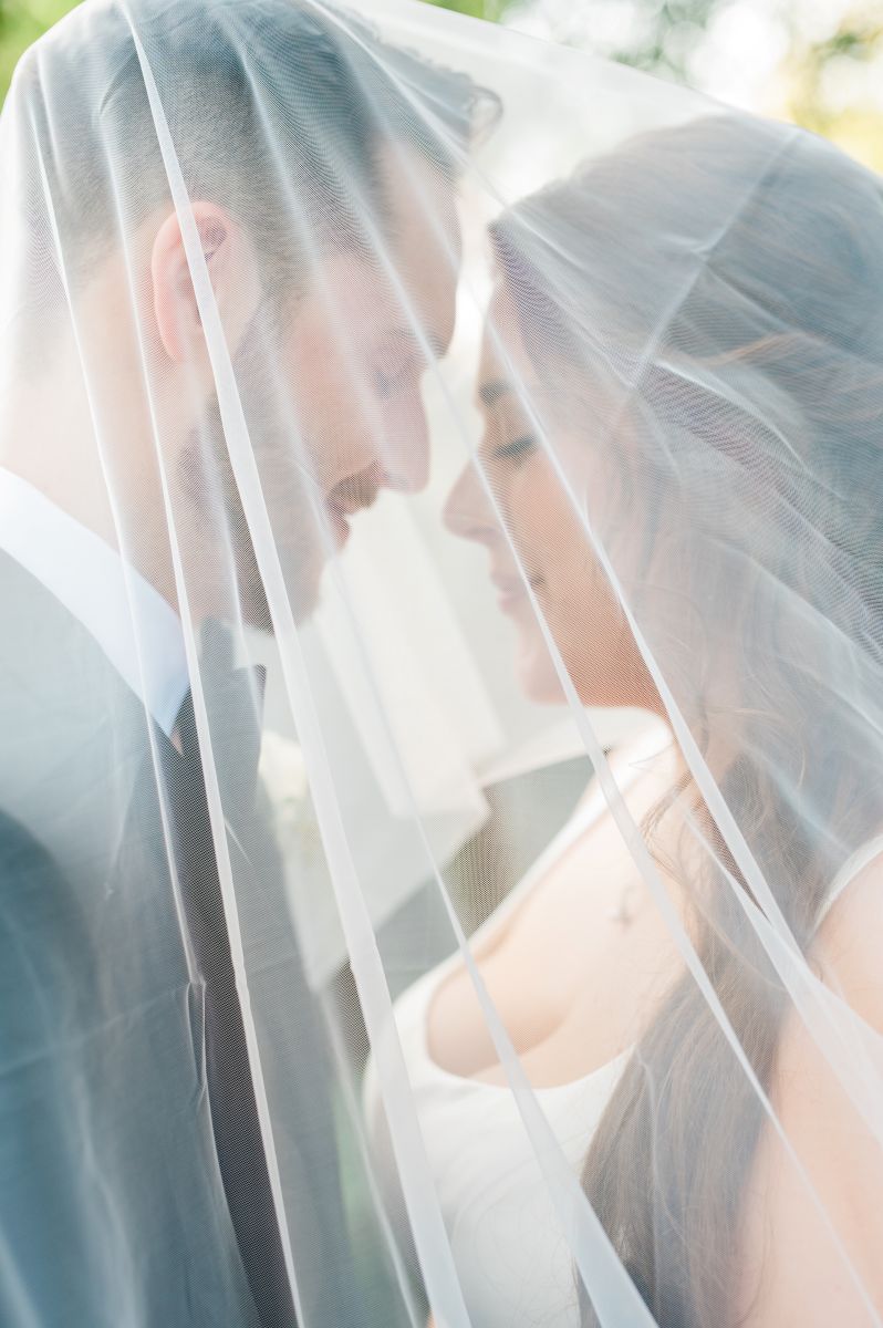 a couple underneath a wedding veil, smiling at each other with their eyes closed the woman is wearing a white wedding dress the man is wearing a black suit