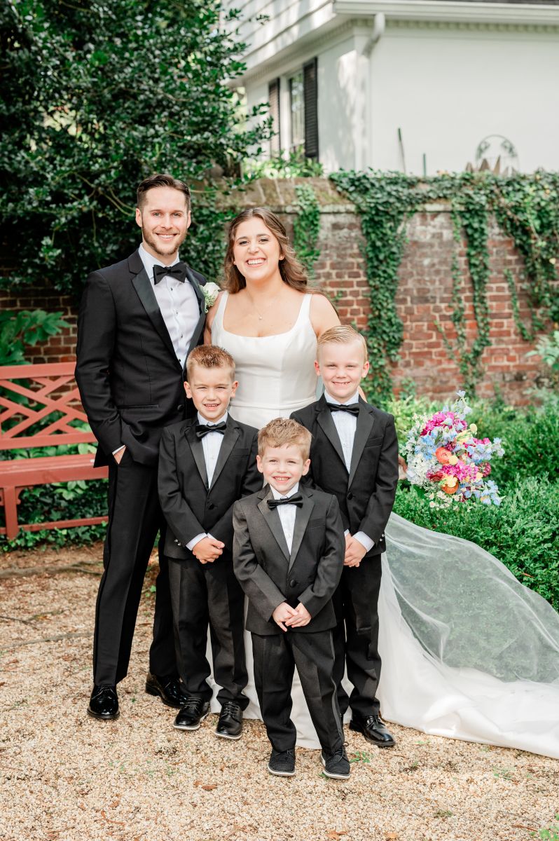 a couple posing in front of a garden and bench with three young boys the boys and man are all wearing black suits the woman is wearing a white wedding dress