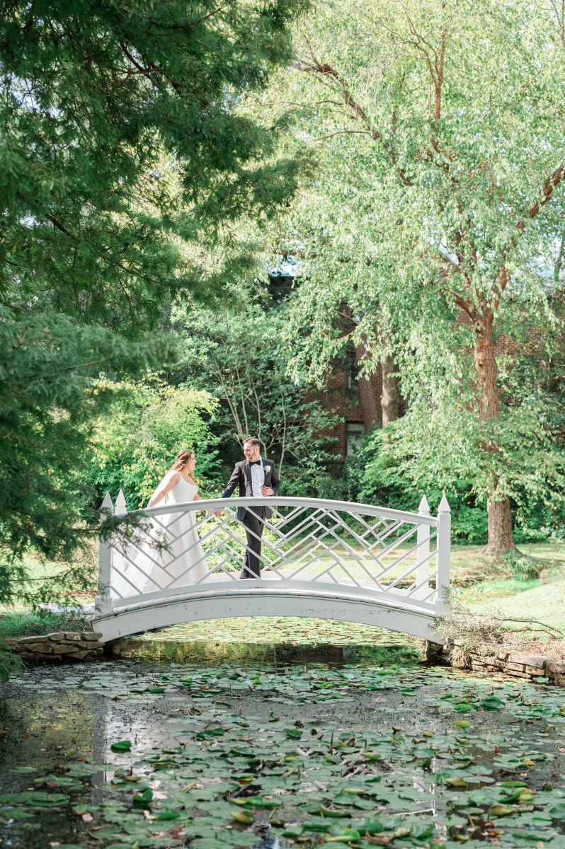 a couple walking across a white bridge next to trees and a pond they are holding hands and looking at each other the woman is wearing a white wedding dress the man is wearing a black suit