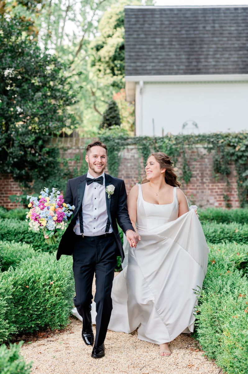 a couple walking through the gradens at william paca house and garden for their wedding the woman is wearing a white wedding dress the man is wearing a black suit and holding a bouquet of flowers