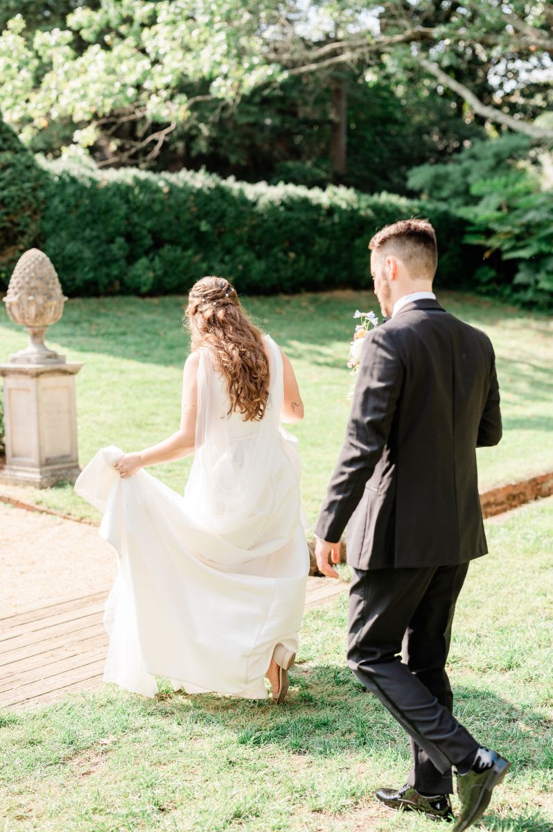a woman is a white wedding dress walking towards a paved path in a garden and a man in a black suit is following her