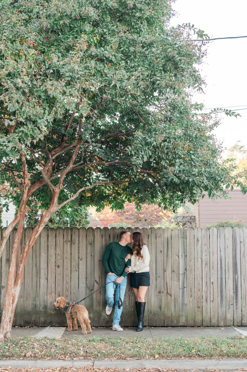 a couple leaning against a wooden fence kissing and holding their dog on a leash