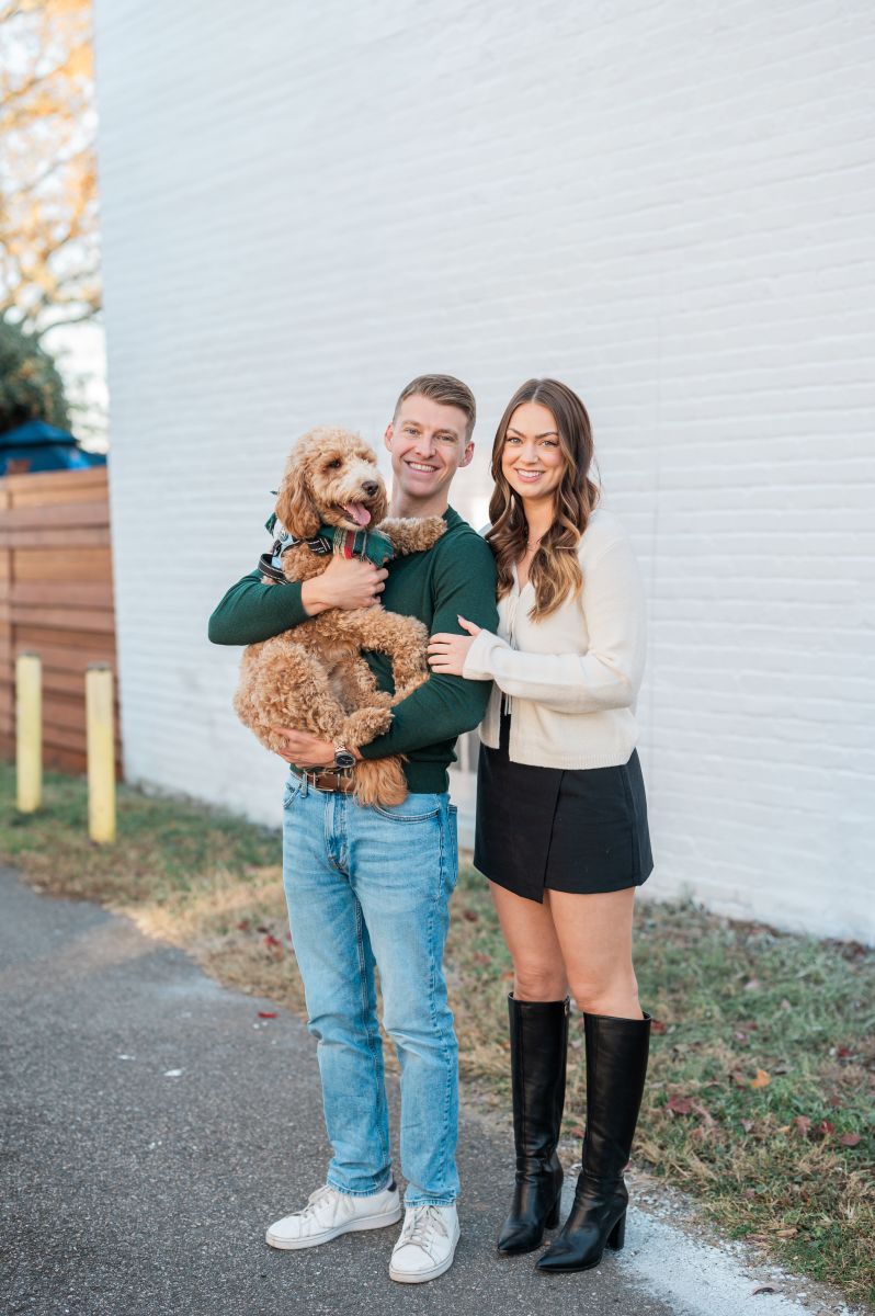 a couple posing in front of a white brick backdrop with their dog