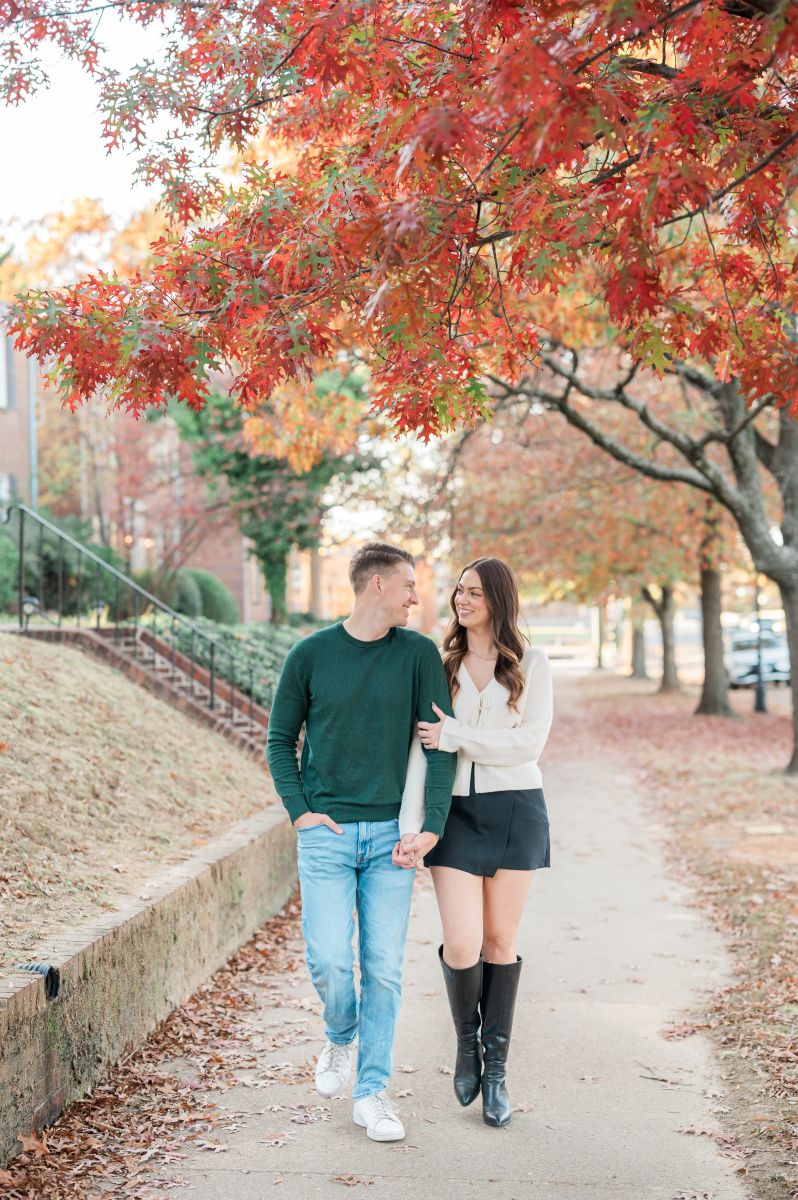 a couple walking down monument avenue in richmond virginia for an engagement session in the fall thye are walking arm in arm and smiling at each other