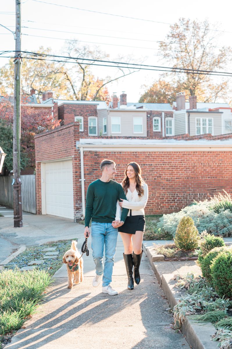 a couple walking together with their dog the couple is holding hands and smiling at each other they are walking on a paved sidewalk