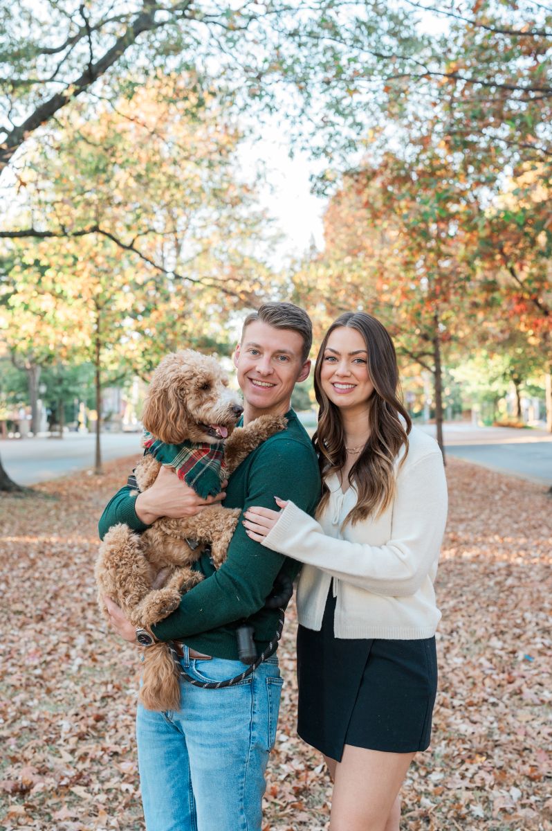 a couple holding their dog on monument avenue in richmond virginia they are surrounded by trees and fall foliage