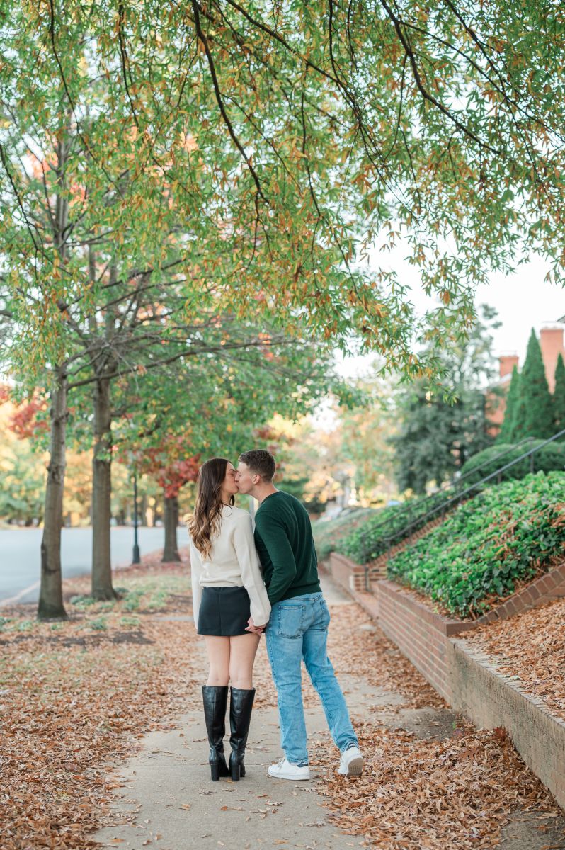 a couple kissin geach other on a sidewalk they are surrounded by fall leaves, trees, and houses