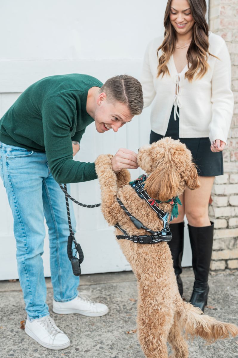 a man giving his dog a treat while his partner stands next to him smiling at them both