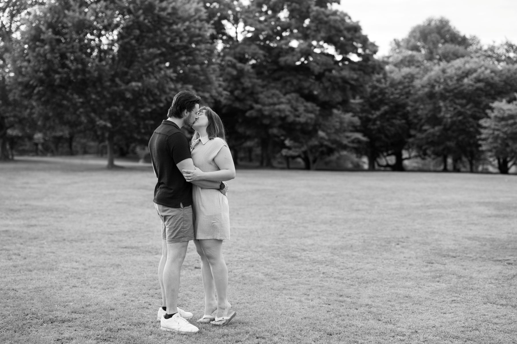 a couple kissing each other and hugging one another on a lawn surrounded by trees