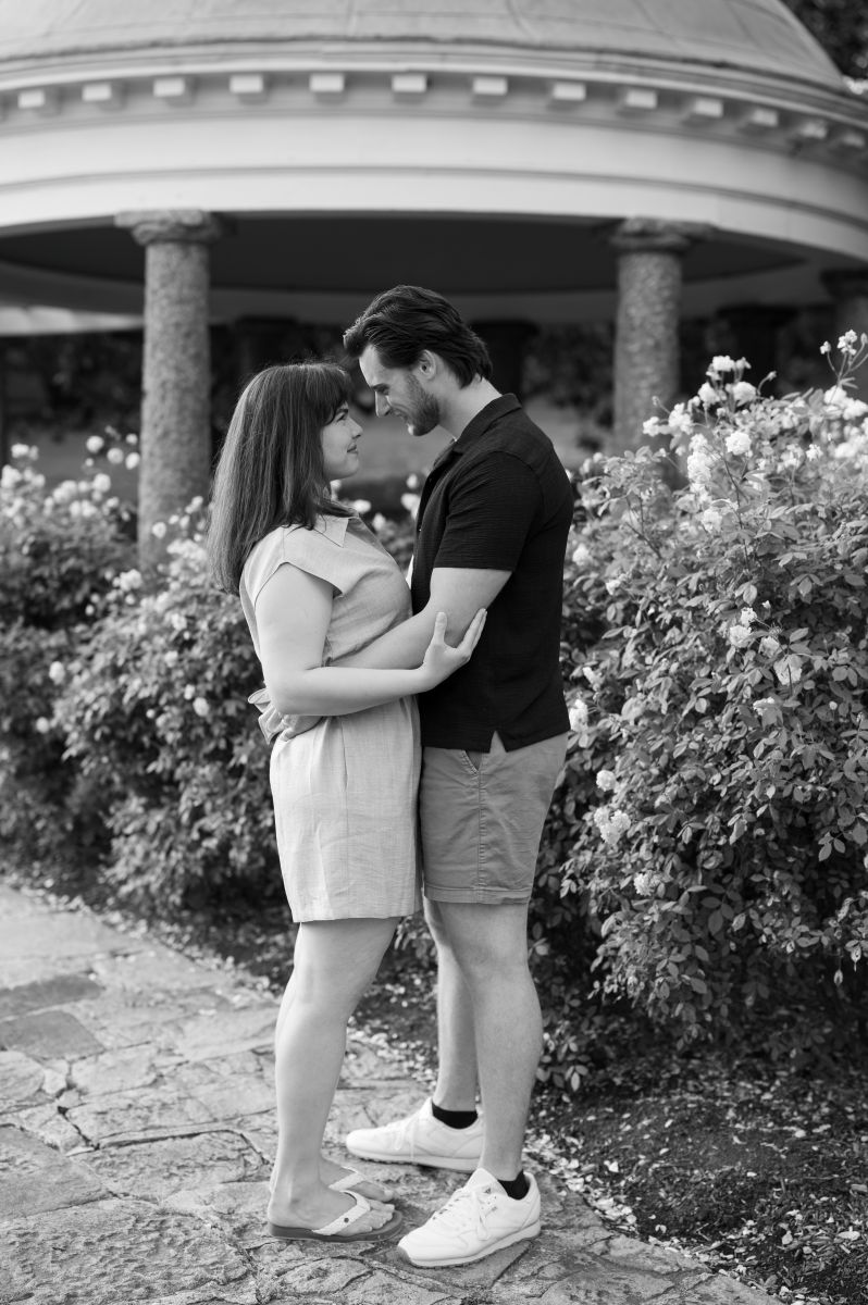 a couple embracing each other and smiling they are next to bushes with flowers and a pavilion