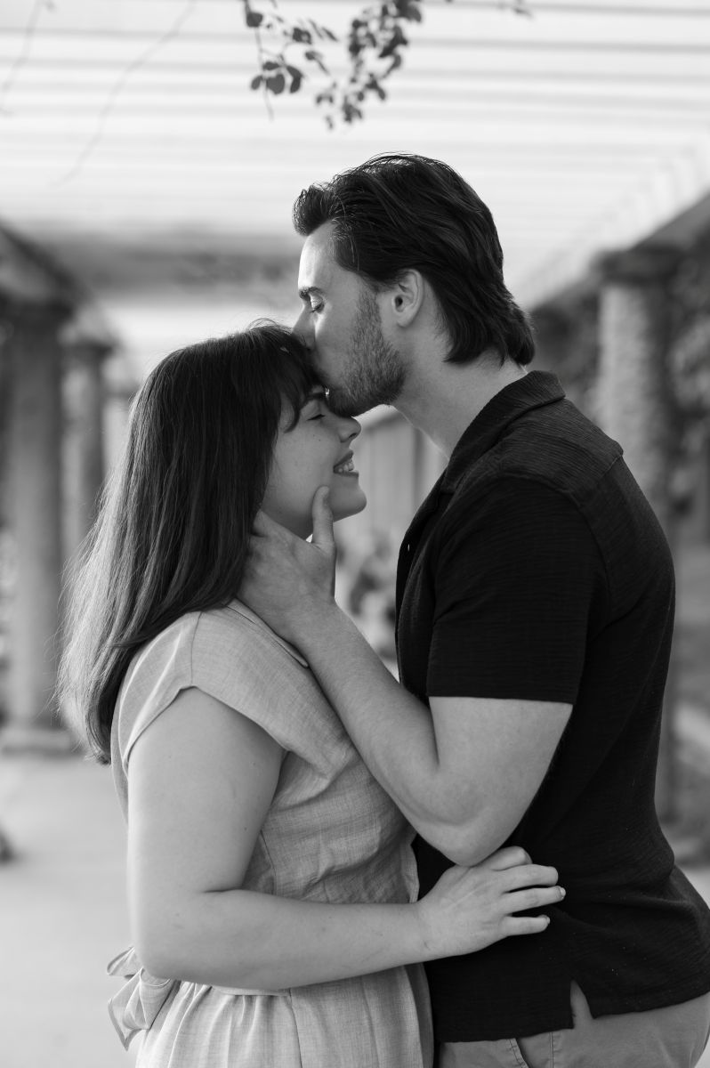 a man with his hands on the side of his partner's neck, leaning in to kiss her on the forehead