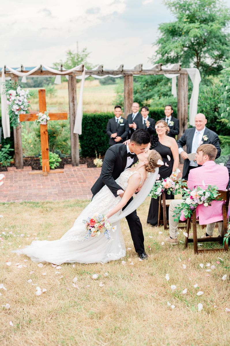 a couple having their wedding at puesta del sol farm in virginia the man is wearing a black suit and dipping his partner who is wearing a white wedding dress and veil backwards as their guests toss white flower petals
