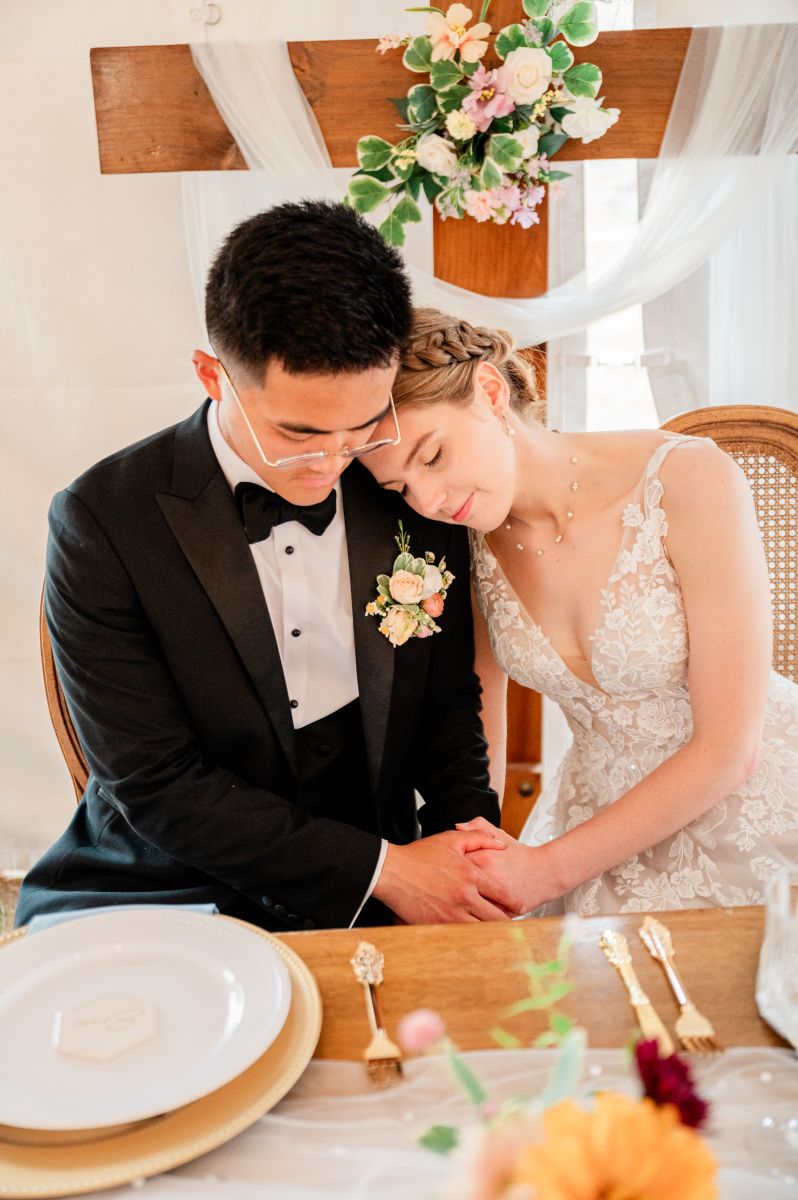 a couple embracing at their wedding table the woman has her head on her partner's shoulder they are both holding hands