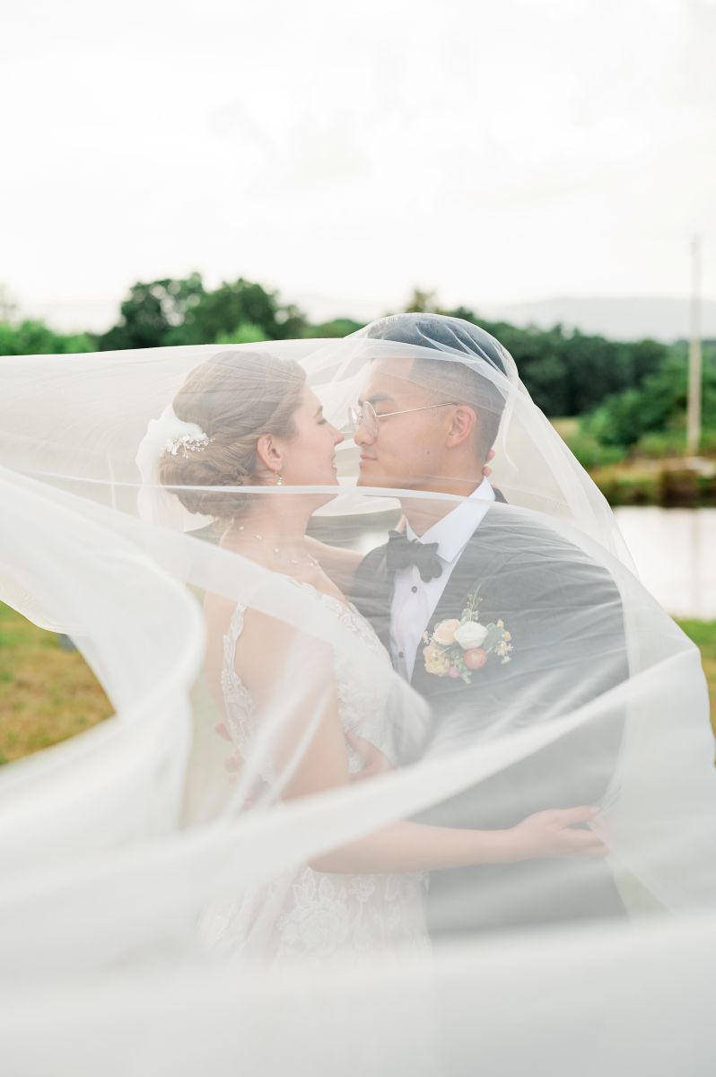 a couple embracing each other underneath the bride's veil she is wearing a white wedding dress and her partner is wearing glasses and a black suit
