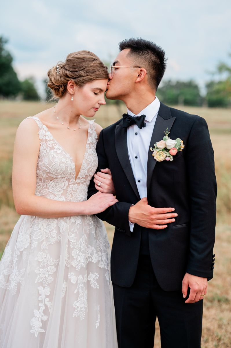 a man wearing a black suit kissing his partner on the forehead next to a field she is wearing a white wedding dress